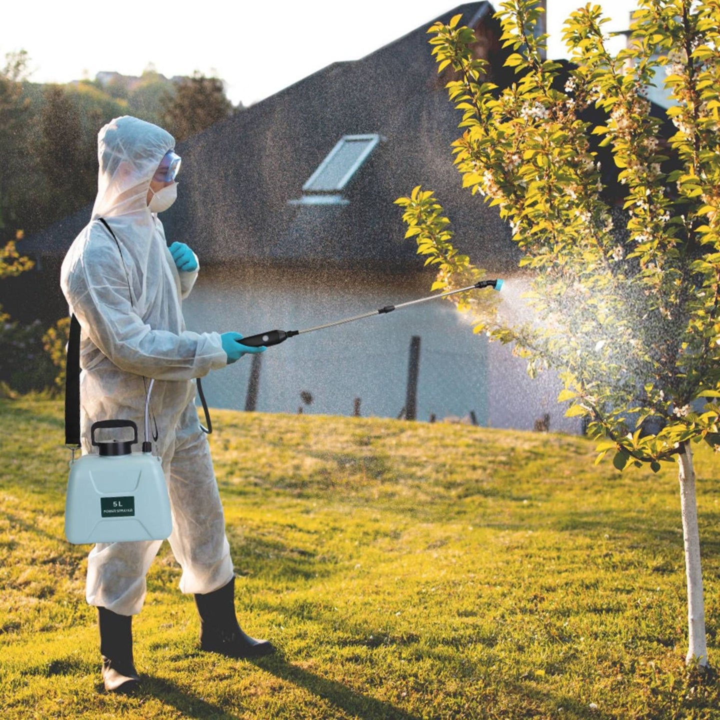 Person in protective suit and mask using an electric sprayer on a tree outdoors for disinfection